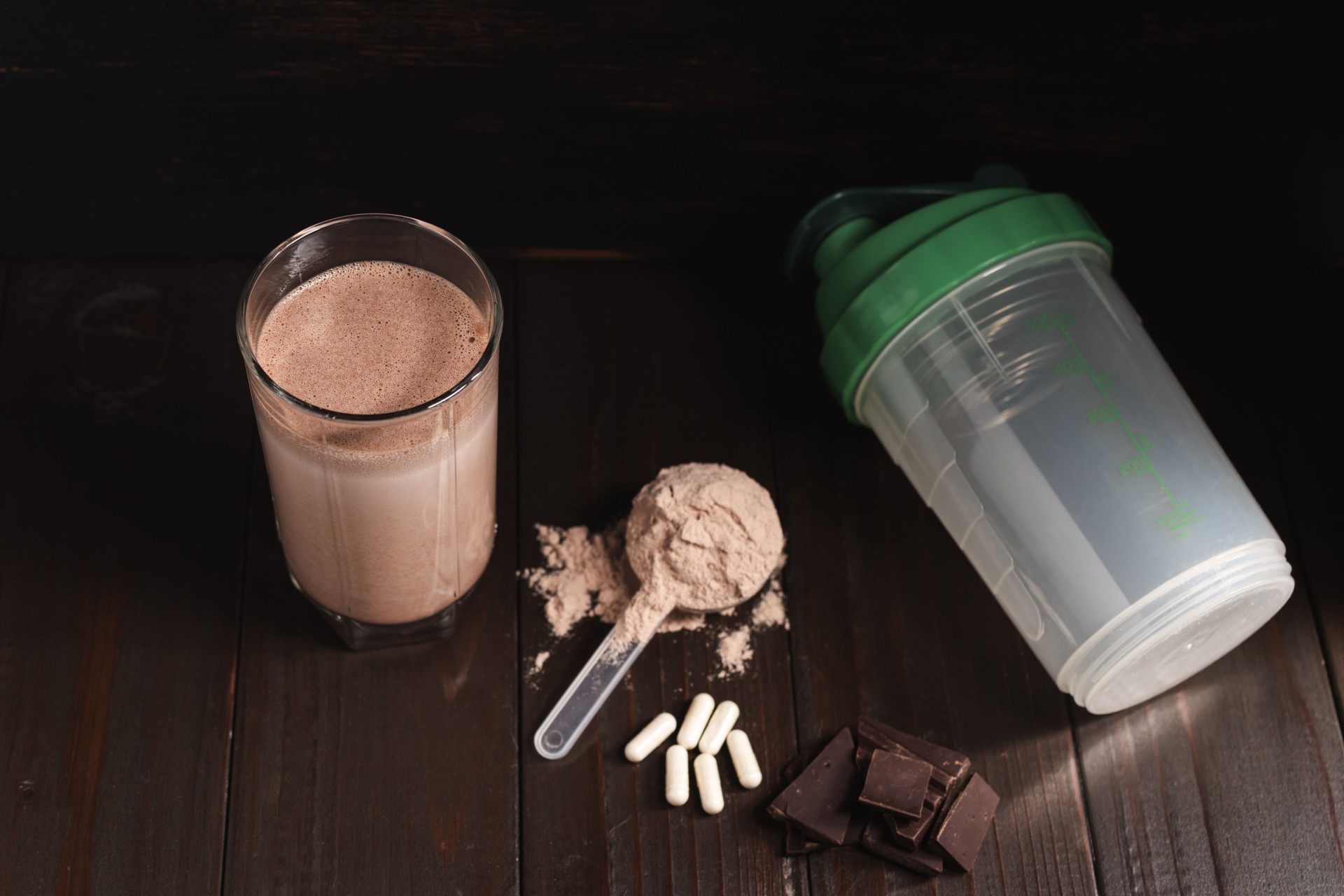 Protein milkshake cocktail in a glass, plastic measuring spoon with whey protein powder, white pills or capsules of amino acids, chocolate cubes on a dark wooden background